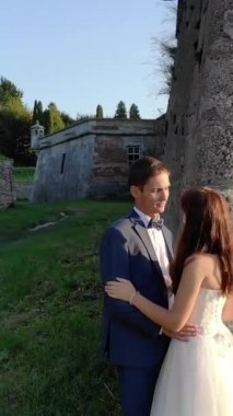 Gelin taş bir duvarın yanında duruyor, gün batımında bir çiftin öpüşmesini izliyor. Yumuşak altın ışık, romantik ve huzurlu bir düğün anında siluetini ön plana çıkarır..