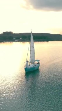 Gün batımında açık denizde seyreden tek bir yatın hava görüntüsü. Dron yukarıdan yelkenliyi yakalarken altın güneş sakin sudan yansıyor..