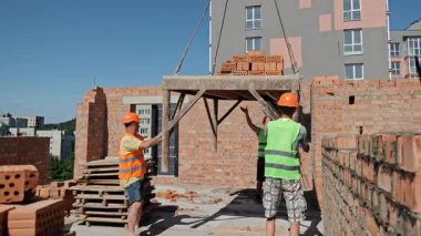 İnşaatçılar iskeleye asılmış tuğlaları yönlendiriyor. Güvenlik yelekleri ve miğferlerle inşaat işçileri vinçle kaldırılan tuğla paletini bir inşaat alanındaki iskeleye yönlendiriyor.