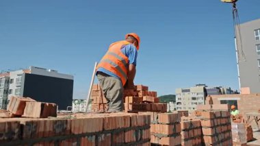 İnşaat işçisi çatı duvarına tuğla diziyor. Şehir inşaatı sırasında açık mavi bir gökyüzünün altında yüksek duvarda içi boş tuğlalar düzenleyen çelik yelek ve miğfer yapımcısı.