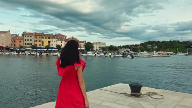 Rıhtımda Yürüyen Kırmızı Elbiseli Kadın Bot ve Renkli Tarihi Binalarla Limana Bakıyor, Rovinj Sahil Kasabasında Yaz Gezisi Konsepti Hırvatistan.