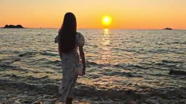 Gün batımında Rovinj Hırvatistan kıyılarında duran kadın, altın güneşin suya yansımasını, gökyüzündeki parlak renklerle romantik deniz manzaralı anı izliyor.