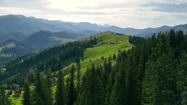 Orman ve dağınık kulübelerle birlikte dağlık dağ sırtı. Kabinler ve yoğun çam ormanları ile benekli yemyeşil dağ sırtı, katmanlı mavi tepeler ve bulutlu bir gökyüzü zeminine karşı kurulmuş.