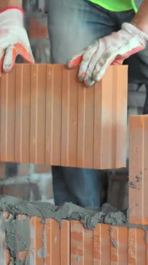 Duvara tuğla ören inşaat işçisi eldivenli bir inşaat işçisi ve güvenlik yeleği kısmen inşa edilmiş bir duvara büyük bir tuğla yerleştiriyor.