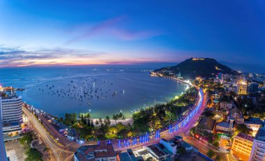 Vung Tau city aerial view with beautiful sunset and so many boats. Panoramic coastal Vung Tau view from above, with waves, coastline, streets, coconut trees and Tao Phung mountain in Vietnam.