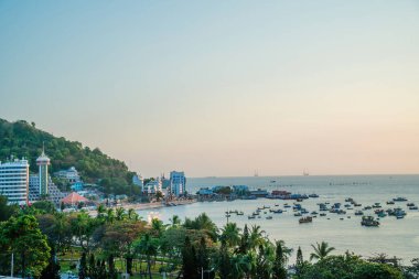 Vung Tau city aerial view with beautiful sunset and so many boats. Panoramic coastal Vung Tau view from above, with waves, coastline, streets, coconut trees and Tao Phung mountain in Vietnam.