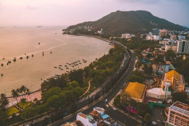 Vung Tau city aerial view with beautiful sunset and so many boats. Panoramic coastal Vung Tau view from above, with waves, coastline, streets, coconut trees and Tao Phung mountain in Vietnam.