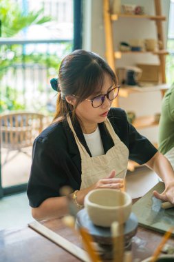 Beautiful woman potter working on potters wheel making ceramic pot from clay in pottery workshop. Focus hand young woman attaching clay product part to future ceramic product. Pottery workshop.