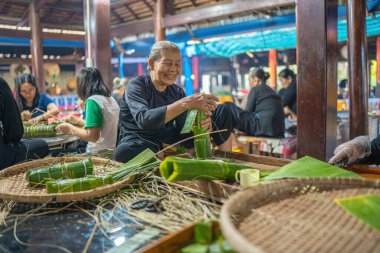 Vung Tau, VIETNAM - JAN 12 2023: Focus old woman with traditional Vietnamese dress (ao ba ba) Making (wrapping) Tet Cake, the Vietnamese lunar new year Tet food outdoor by hands.