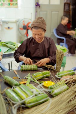 Vung Tau, VIETNAM JAN 20 2023: Focus old Buddhist nun with traditional Vietnamese dress (ao ba ba) Making (wrapping) Tet Cake, the Vietnamese lunar new year Tet food outdoor by hands.