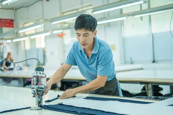 Ba Ria, VIETNAM - JAN 19 2023: Man with cutter machine and personal protective equipment at garment industrial work place. Fabric cutter in Asian textile garment factory