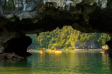 Ha Long Körfezi 'ndeki kireçtaşı kayalarının arasında yüzen turist hurdaları, Halong Körfezi, Vietnam yakınlarındaki Lan Ha Körfezi' ndeki bir mağarada kano yapan insanlar.