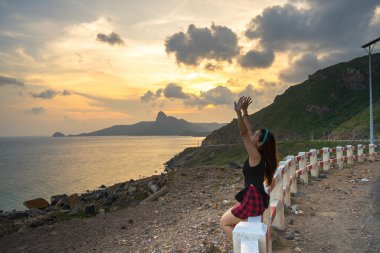 Con Dao adası kıyı manzarasının önünde oturan genç turistin arkası. Dalgalar, kıyı şeridi, açık gökyüzü ve yol, Bai Nhac plajında mavi deniz ve dağ.