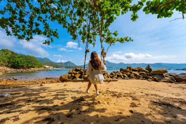 Sahilde salıncakta sallanan seyahat eden kadının arka planında palmiye ağaçları, Hint badem ağacı ve turkuaz deniz var. Yaz tatili, tropik tatiller. Seyahat ve yaşam tarzı kavramı.
