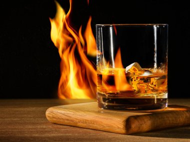 Glass with whisky with ice on wooden table and black background with flame
