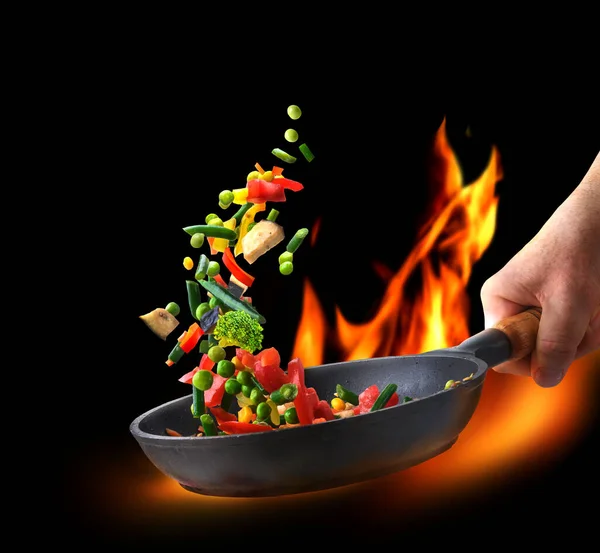 Various chopped vegetables flying into a pan in chef hand on flame and black background