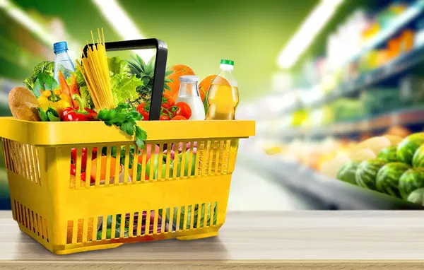 Food and groceries in shopping basket on wood table on blurred suppermarket aisle background