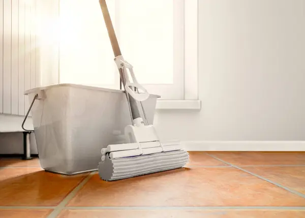A mop and bucket are on the kitchen floor, wet cleaning. The tiled floor is made of composite material