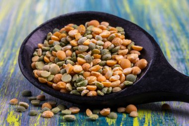 Ahşap bir kaşık, parlak mavi ve yeşil bir zemin karşısında, sarı ve yeşil bezelyeler de dahil olmak üzere renkli mercimeklerden oluşan bir karışım barındırır. Mercimek çeşitli boyutlarda ve dokularda sergilenir.