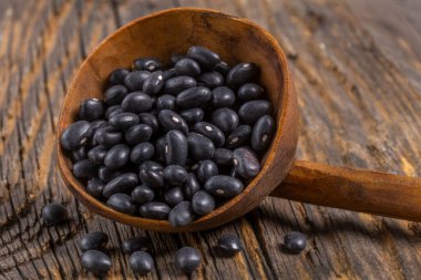 Ahşap bir kaşık, kaba ahşap bir yüzeyin zemininde duran parlak siyah fasulyelerle doludur. Fasulyeler parlak bir doku sergiliyor, ışığı yansıtıyor ve koyu renklerini sergiliyorlar.