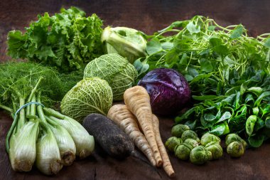 Yapraklı yeşillikler, lahanalar, kök sebzeler ve otlar da dahil olmak üzere çeşitli taze sebzeler, canlı renkler ve dokular içeren, kırsal ahşap bir yüzey üzerinde düzgünce düzenlenmiştir..