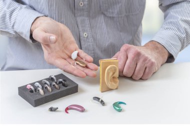 Masada farklı tip işitme cihazları ve kulak modelleri var. Diğer cihazlar yakınlarda gösterilirken birey bir aygıtı elinde tutuyor, işitme yardımı seçeneklerini vurguluyor.