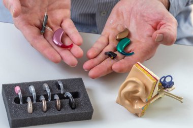 Ellerinde çeşitli işitme cihazları ve yardımcı cihazlar var, farklı stil ve renkler sergiliyorlar. Bir model kulak masaya oturur ve işitme güçlendirici teknolojiyi vurgular..