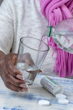 Pembe eşarplı bir insan, şişeden ahşap bir masaya dinlenen temiz bir bardağa su döker. Yakınlarda beyaz tabletler ve bir konteynır var. Bu da sağlık durumunun normal olduğunu gösteriyor..