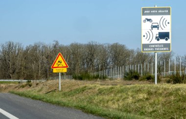 Trafik işaretleri sürücülere otoyol kenarındaki hız kontrolü için sık sık radar kontrolü yapmalarını bildiriyor. Bölge kırsal, arka planda açık bir manzara var..