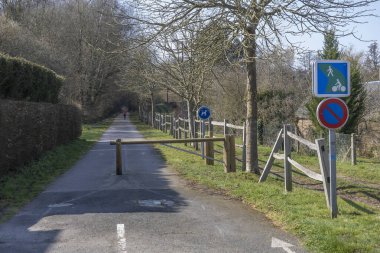 Ağaçlar ve yeşilliklerle çevrili bir bisiklet yolunu engelleyen ahşap bir bariyer. Uzaktan bir yaya görülebiliyor ve işaretler yürüyüş ve bisiklet alanlarını gösteriyor..