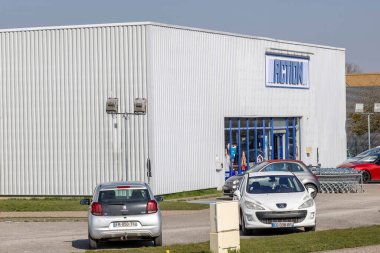 Geniş park alanı olan bir perakende mağazasında aydınlık bir gün. Birkaç araba park edilmiş ve alışveriş arabaları girişin dışında düzenli bir şekilde düzenlenmiş. Müşterileri alışveriş deneyimi için karşılıyorlar..