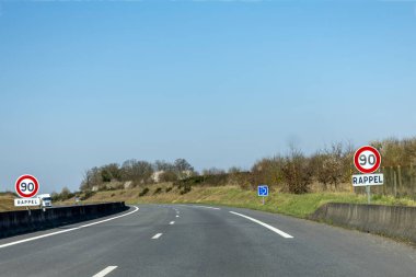 İki hız limiti işareti kırsal bir yolda maksimum 90 km hız olduğunu gösteriyor. Güneşli hava ve açık mavi gökyüzü kırsal alandaki sakin manzarayı güçlendirir..