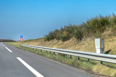 Hız limiti işareti ve yemyeşil otların arasında yön işareti gösteren bir otoyol manzarası. Gökyüzü açık mavi, güneşli bir günü işaret ediyor..