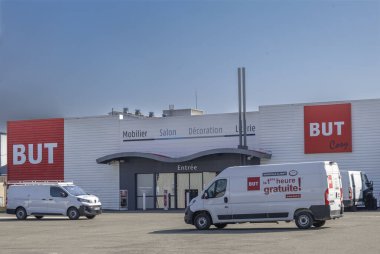 Yoğun bir mobilya mağazası büyük tabelaları olan modern bir dış görünüşü sergiliyor. Teslimat araçları dışarıda park halinde. Ticari bir bölgede faaliyet günü olduğunu gösteriyor..