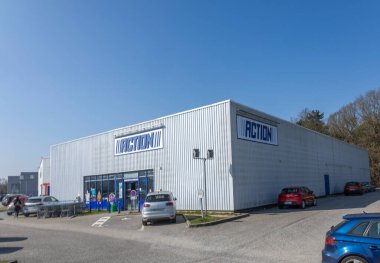 Açık mavi gökyüzünün altında hareketli bir indirim mağazası görülebilir. Binanın etrafına park edilmiş arabalar, gün içinde aktif müşteri ziyaretleri olduğunu gösteriyor..