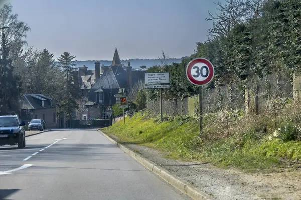 Virajlı bir yol büyüleyici bir kır evine götürür. 30 km 'lik hız limiti işareti görülebiliyor, bu da sakin bir alanı gösteriyor. Yeşillik yemyeşil çizgiler açık gökyüzünün altında yol kenarı.