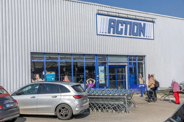 Alışverişçiler güneşli bir günde perakende mağazasının önünde toplanırlar. Alışveriş arabaları düzenli bir şekilde yerleştirildi ve araçlar yakınlarda park halinde, hareketli bir atmosfer yaratıyorlar..