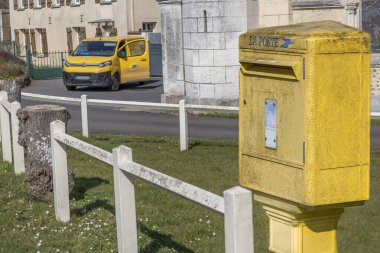 Küçük bir kasabada sakin bir cadde boyunca bir posta arabası yaklaşırken canlı sarı bir posta kutusu beyaz bir çitin yanında duruyor. Sahne, günlük hayatı huzurlu bir ortamda yakalıyor..