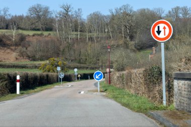 Kırsal kesimde bir kır yolunda trafik işaretleri yol gösteren araçlar bulunur. İşaretler tek yönlü ve çift yönlü trafiği gösterirken yeşil alan açık mavi gökyüzünü gösteriyor..