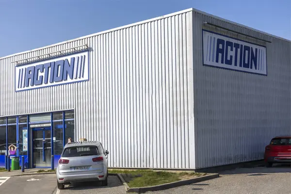 Ticari bir alanda büyük bir perakende binası tepede parlak mavi bir gökyüzüne sahiptir. Arabalar dışarıda park halinde ve mağaza levhası belirgin bir şekilde sergileniyor. Müşteriler içeri davet ediliyor..