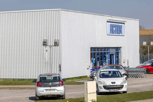 Geniş park alanı olan bir perakende mağazasında aydınlık bir gün. Birkaç araba park edilmiş ve alışveriş arabaları girişin dışında düzenli bir şekilde düzenlenmiş. Müşterileri alışveriş deneyimi için karşılıyorlar..