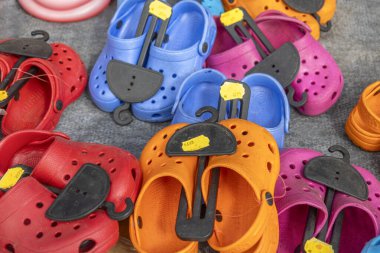 Bir pazarda çeşitli renkli takunyalar düzenlenir. Ayakkabılar kırmızı, mavi, turuncu ve pembe tonlarını içeriyor, her biri canlı bir seçim sergileyerek fiyat etiketleriyle etiketlenmiş..