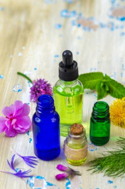 Temel yağları içeren çeşitli renkli şişeler taze bitki ve çiçeklerin yanı sıra ahşap bir yüzey üzerinde düzenlenmiştir. Bu da doğal ilaçlara ve aromaterapiye odaklanmayı önerir..