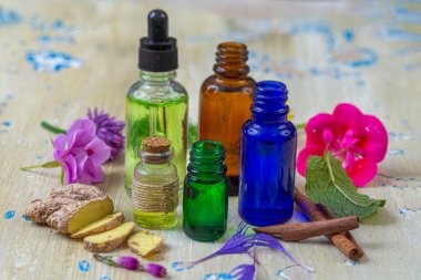 Tahta bir masanın üzerinde zencefil, çiçek ve tarçın ile birlikte farklı renklerde çeşitli yağlar sergilenerek, doğal malzemelerin canlı bir düzenini yakalar..