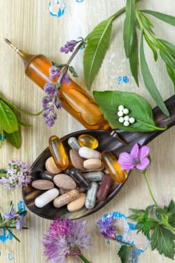 Vitamin çeşitleri, bitkisel kapsüller ve tahta kaşıkta sergilenen küçük bir şişe. Taze bitkiler ve çiçekler kaşığın etrafını sarıp canlı, organik bir ortam oluşturuyor..