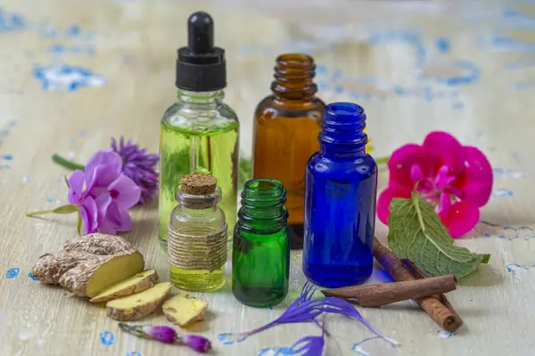 Tahta bir masanın üzerinde zencefil, çiçek ve tarçın ile birlikte farklı renklerde çeşitli yağlar sergilenerek, doğal malzemelerin canlı bir düzenini yakalar..