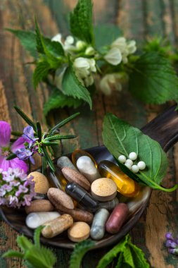 Tahta kaşık, bitkisel ilaçlardaki sağlık ve sağlığa olan doğal yaklaşımı gösteren taze bitkiler ve çiçeklerle çevrili çeşitli bitkisel takviye maddeleri ve vitaminleri barındırır..