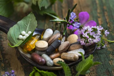 Tahta bir kaşıkta sergilenen çeşitli bitkisel takviyeler ve vitaminler. Taze otlar ve rengarenk çiçekler kaşığın etrafını sarıp, doğal sağlık ve refahı ön plana çıkarıyor..