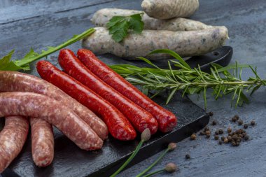 Siyah bir kesme tahtasında farklı türde sosisler sergilenir. Biberiye ve soğan gibi taze otlar canlı renkler ekler ve lezzetli bir yemek hazırlığını güçlendirir..