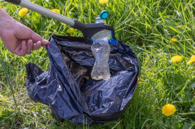 Birisi, sarı karahindiba dolu yemyeşil bir alandan plastik bir şişeyi çıkarmak için çöp toplayıcı kullanıyor. Çaba, çevre temizliği ve farkındalığı teşvik ediyor.